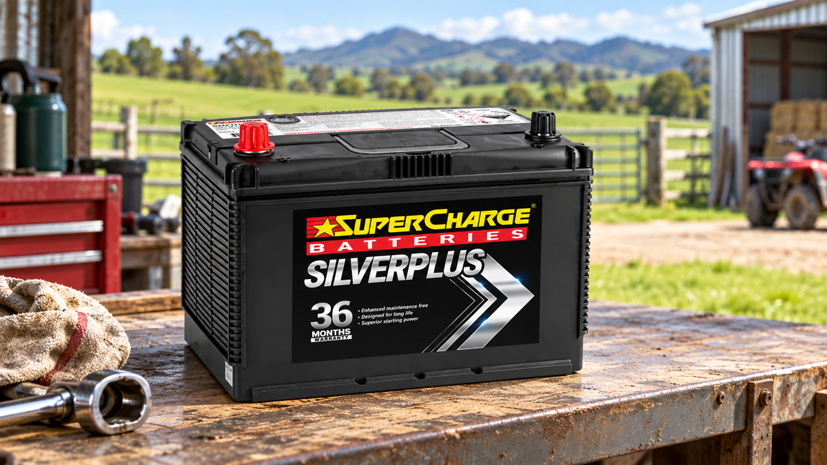 SuperCharge SilverPlus car battery on a rural New Zealand farm workshop bench, with green paddocks, farm sheds and hills in the background.