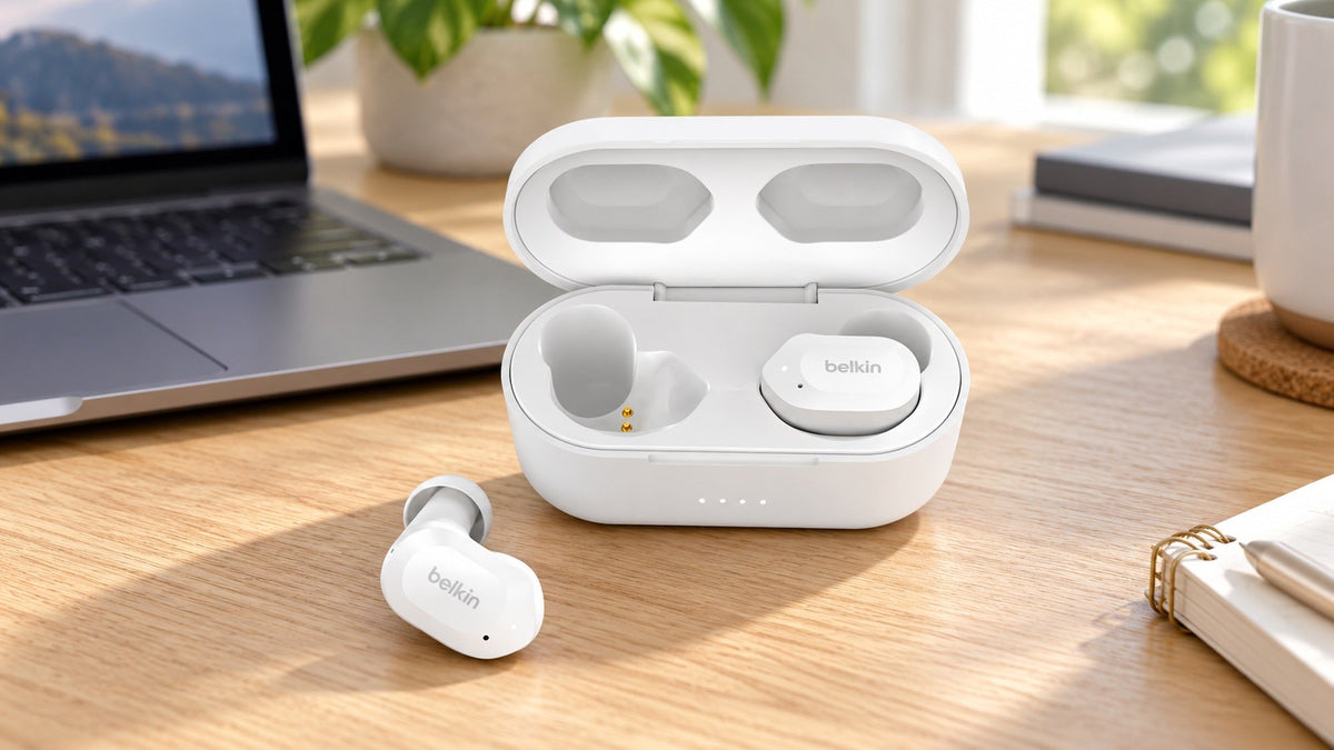 White Belkin wireless earbuds and an open charging case on a wooden desk beside a laptop.