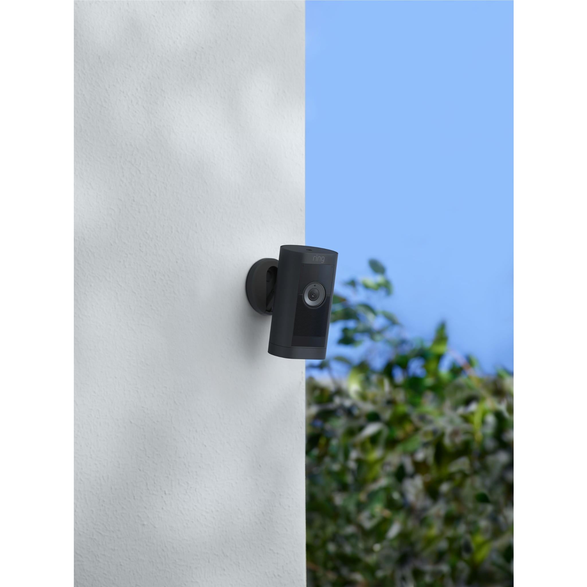Black stick-up security camera mounted on exterior wall near greenery