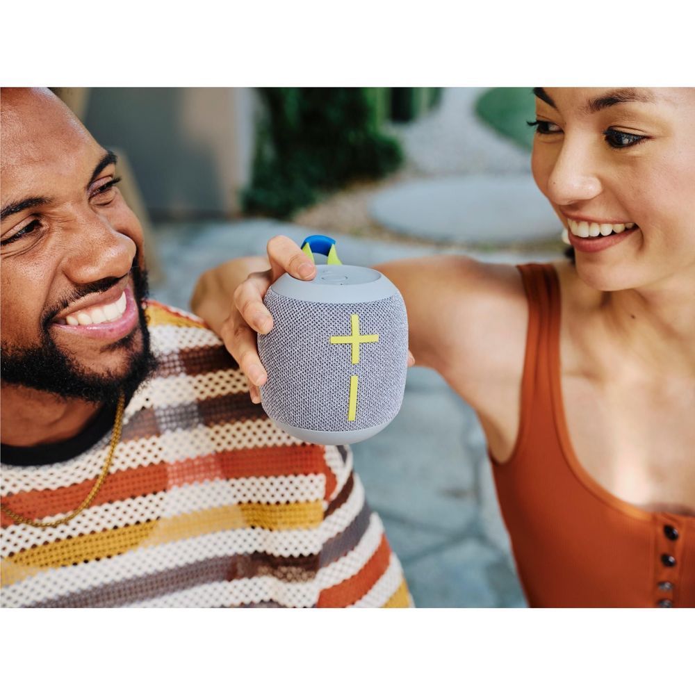 Gray fabric portable Bluetooth speaker with lime strap and yellow plus, held in hands