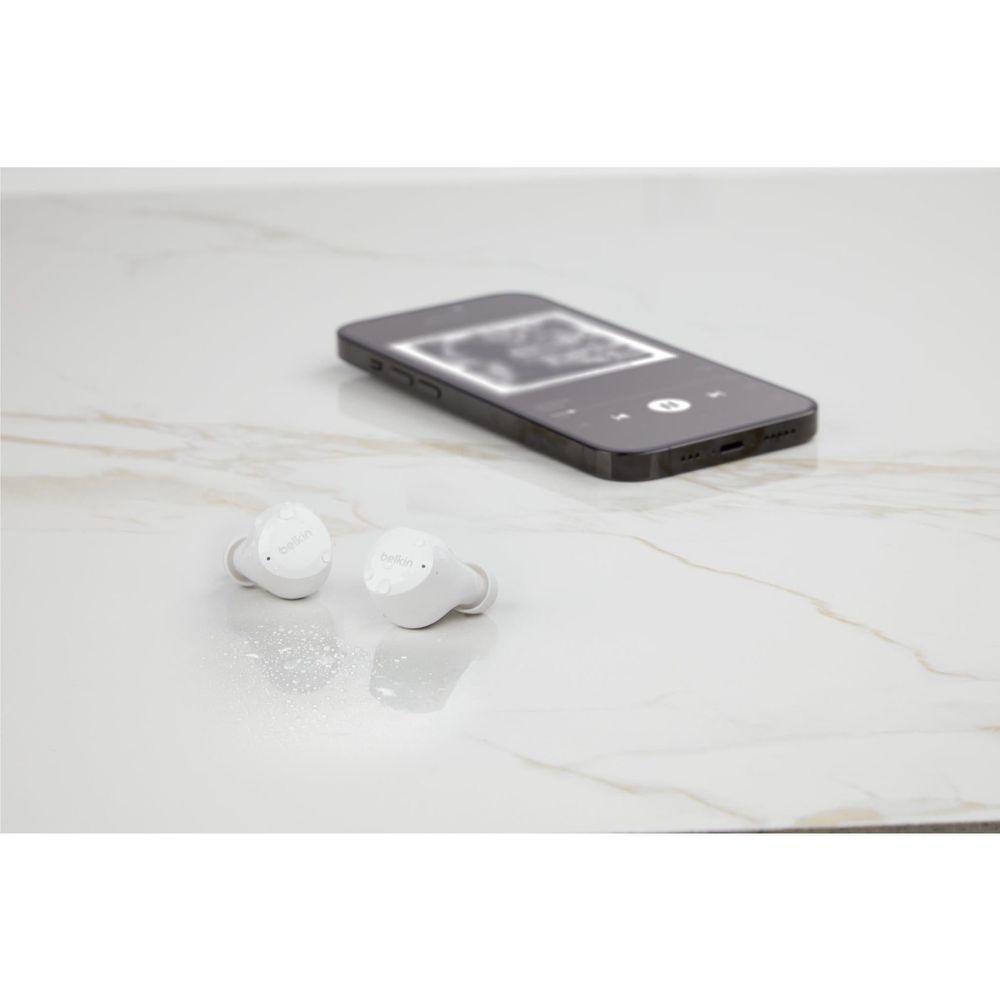 White Belkin earbuds and charging case on a surface near a smartphone