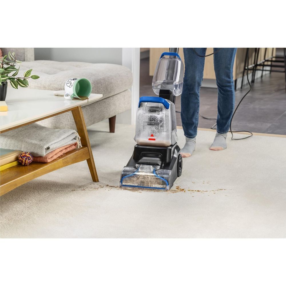 Upright cleaner treating a light rug beside furniture; clear tanks and blue head