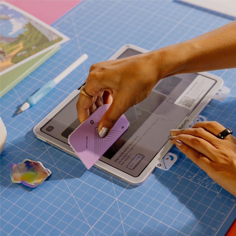Hands applying a purple-tinted screen protector to an iPad Pro on a blue mat