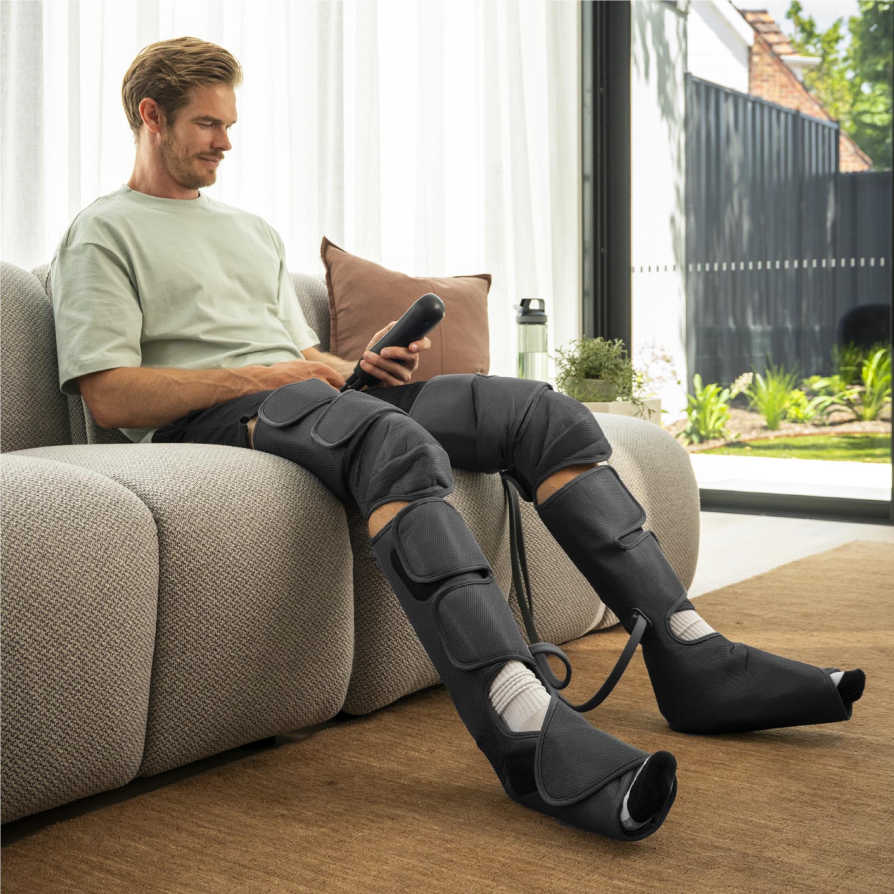Leg compression massager worn by man seated on couch, dark gray wraps visible