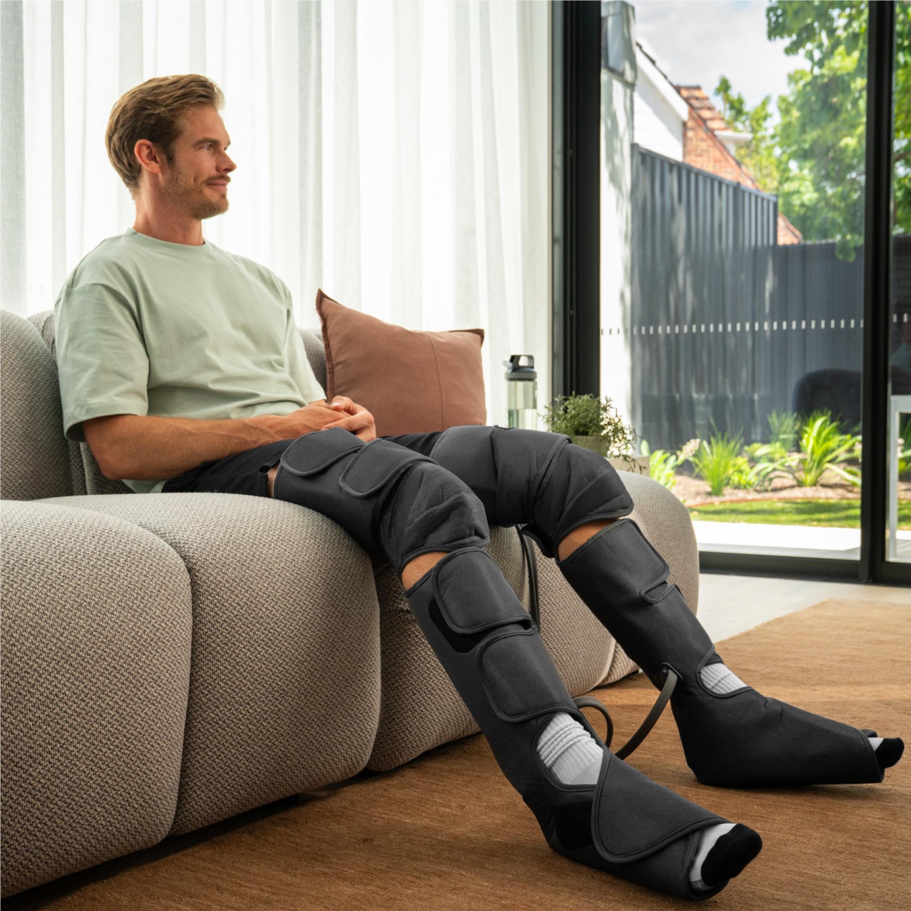 Leg compression massager worn by man reclined on couch, dark gray knee pads