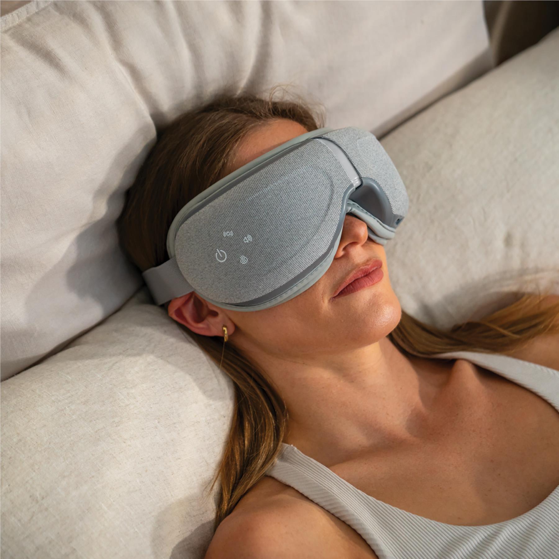 Grey eye massager worn on face with elastic strap, woman reclining on a sofa