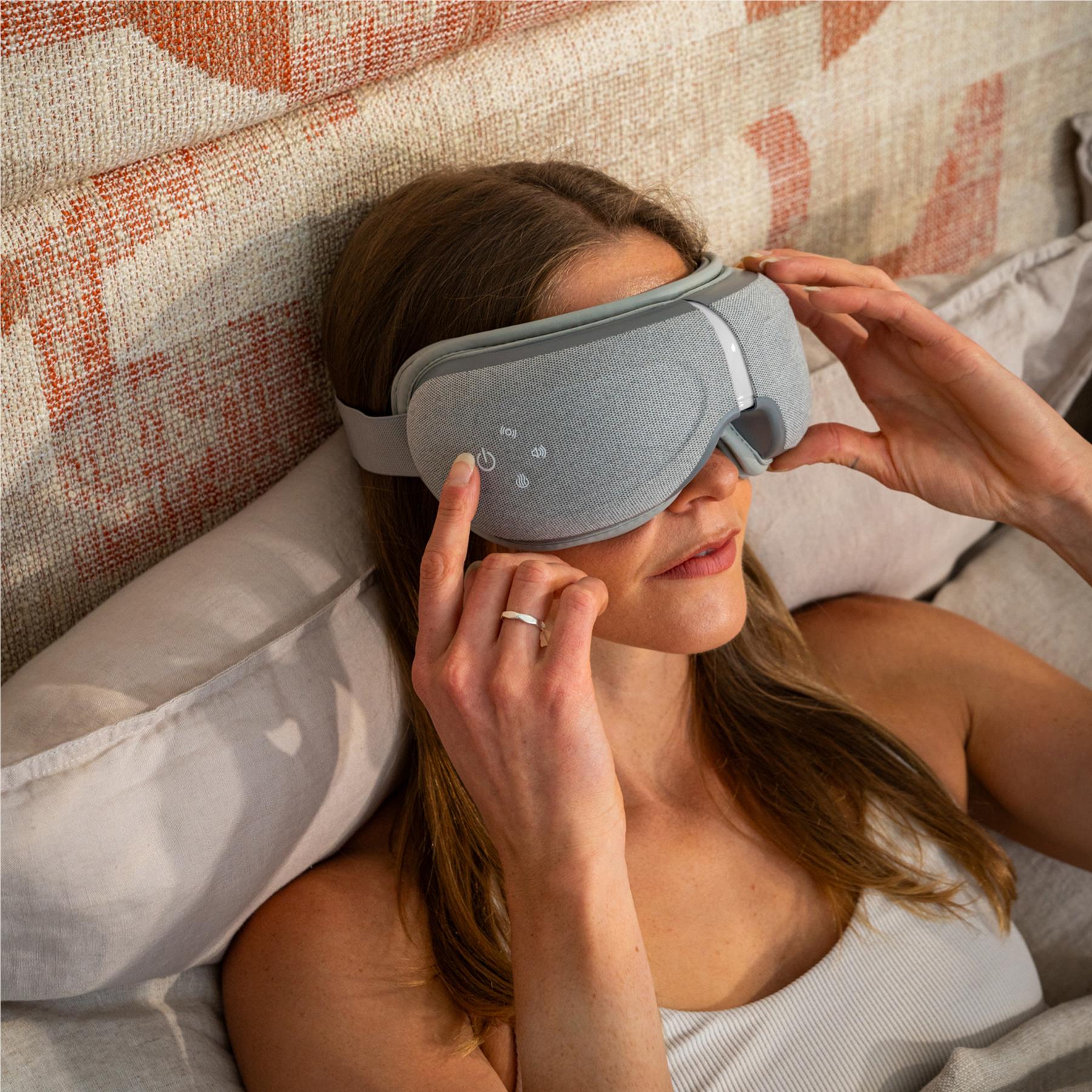 Grey eye massager worn over closed eyes, woman adjusting the elastic head strap