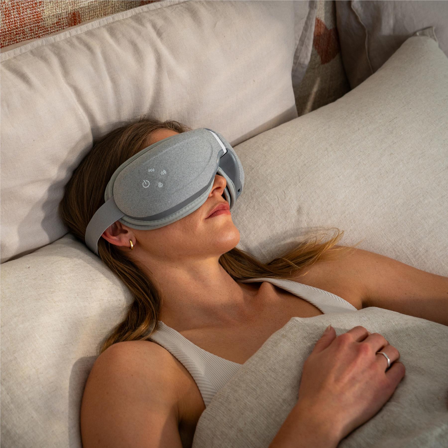Grey eye massager worn on eyes while reclining on a sofa, padded visor and strap