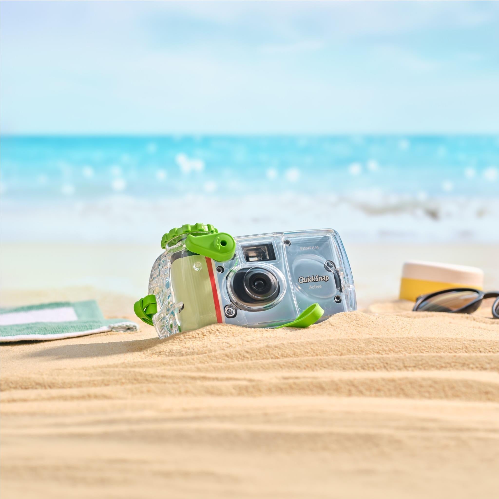 Clear waterproof disposable camera with green accents and red stripe, lying on sand.