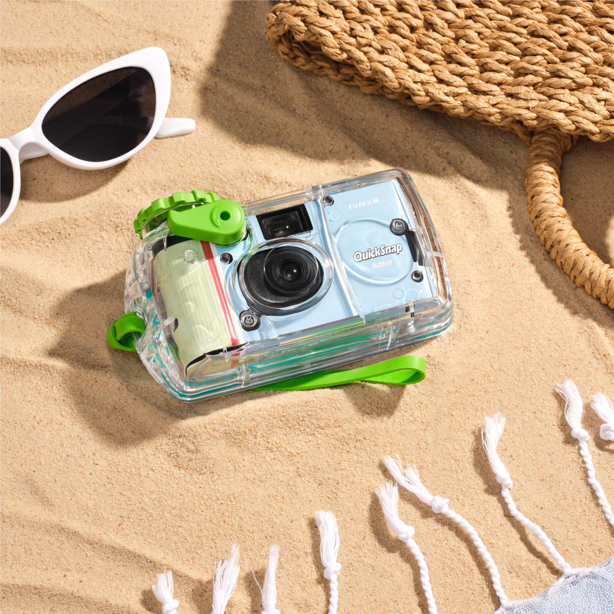 Clear disposable camera with green latch and lens, on sand near seashells.
