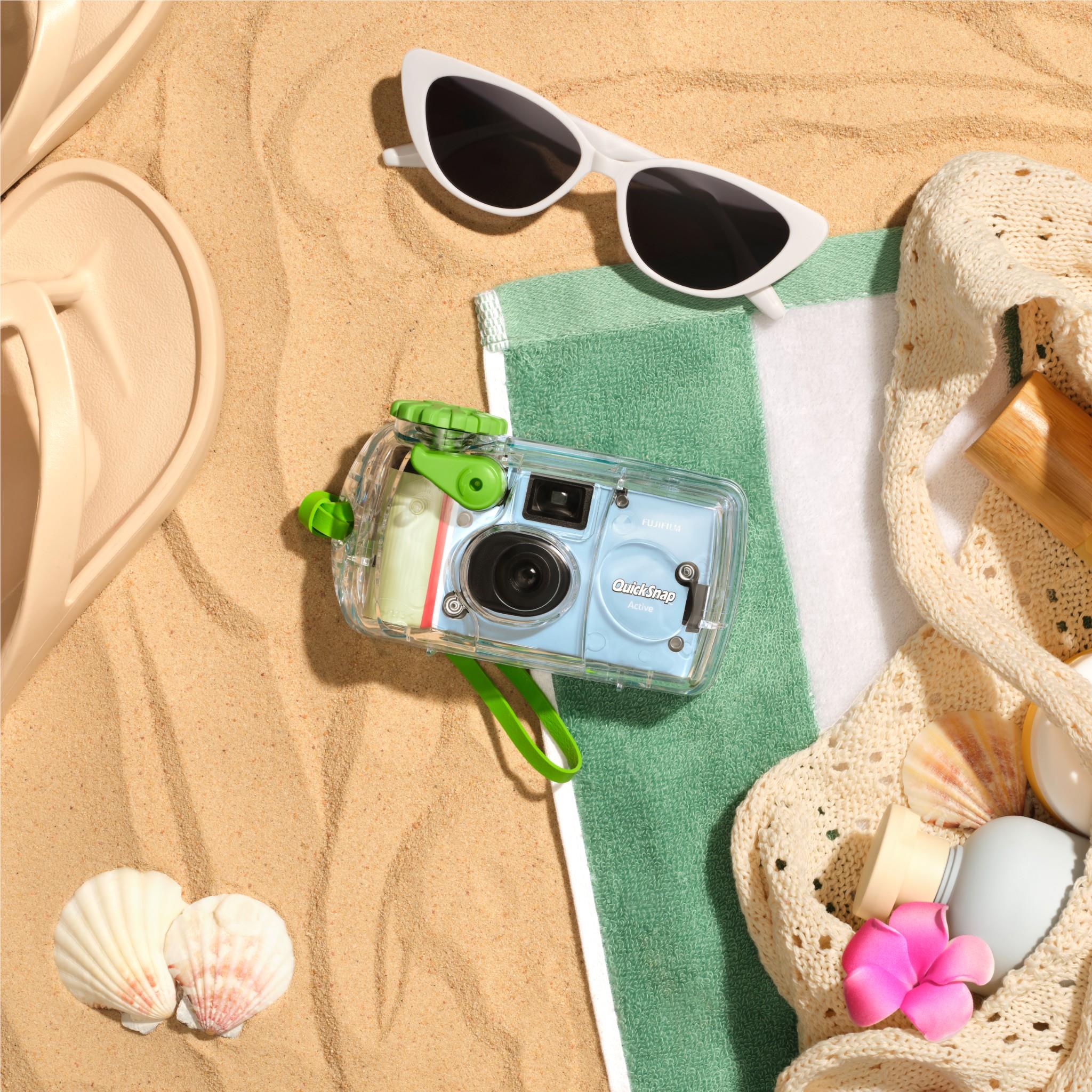 Clear green-accent disposable camera on a towel with sunglasses and seashells.